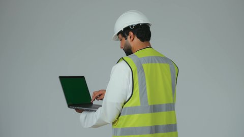 Engineering project management, reliance on modern technology and techniques in the engineering sector, a portrait from behind of an Emirati Gulf Arab engineer wearing a kandura, helmet, and safety vest holding a laptop, monitoring his work remotely, typing on the keyboard, white background.