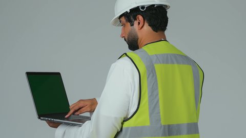 العمل في القطاع الهندسي، الاعتماد على التكنولوجيا والتقنية الحديثة بالقطاع الهندسي، بورتريه مقرب من الخلف لمهندس عربي خليجي إماراتي يرتدي الكندورة والخوذة وسترة الحماية يمسك الحاسوب المحمول يقوم بمتابعة أعماله عن بعد، النقر على لوحة المفاتيح، خلفية بيضاء