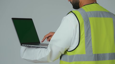 الاعتماد على التكنولوجيا والتقنية الحديثة بالقطاع الهندسي، بورتريه مقرب من الخلف لمهندس عربي خليجي إماراتي يرتدي الكندورة والخوذة وسترة الحماية يمسك الحاسوب المحمول يقوم بمتابعة أعماله عن بعد، النقر على لوحة المفاتيح، خلفية بيضاء