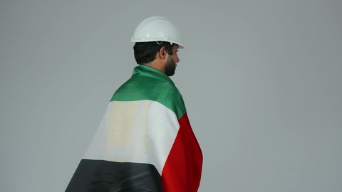 هوية اليوم الوطني الاماراتي 2 ديسمبر، النظر الى الكاميرا بايماءات الاعتزاز والثقة، الحب والانتماء للوطن، احياء ذكرى استقلال الإمارات 1971،  بورتريه لمهندس عربي خليجي إماراتي يرتدي الكندورة والخوذة وسترة الحماية يحمل علم دولة الإمارات المتحدة على ظهره، خلفية بيضاء