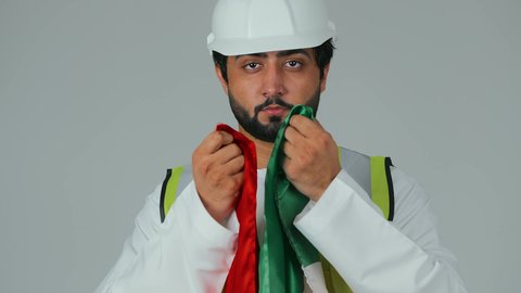 يوم العلم الإماراتي 3 نوفمبر، الاعتزاز بالانتماء للوطن، ذكرى قيام الاتحاد 2 ديسمبر 1971، بورتريه مقرب لمهندس عربي خليجي إماراتي يرتدي الكندورة والخوذة وسترة الحماية يقبل علم دولة الإمارات المتحدة، العمل في القطاع الهندسي، خلفية بيضاء