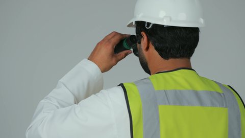 Planning and research for the future, the engineering sector in the UAE, expanding the field of vision, supervising engineering projects, a portrait from behind of an Arab Gulf Emirati engineer wearing a kandura, helmet, and safety vest looking through binoculars, white background.