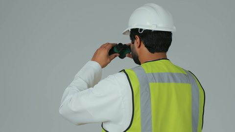 The engineering sector in the UAE, expanding the field of vision, supervising engineering projects, a portrait from behind of an Emirati Gulf Arab engineer wearing a kandura, helmet, and safety vest looking through binoculars, planning and researching for the future, white background.