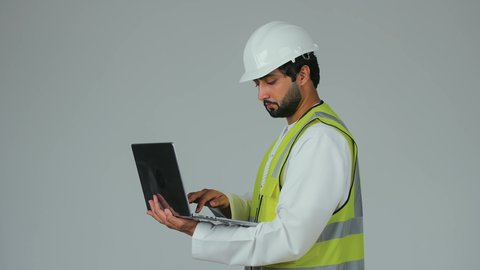 Gestures of integration and focus, completing remote work tasks, typing on the keyboard, using modern technical devices, a portrait of an Emirati Gulf Arab engineer wearing a kandura, helmet, and safety vest using a laptop, following architectural and engineering projects, white background.