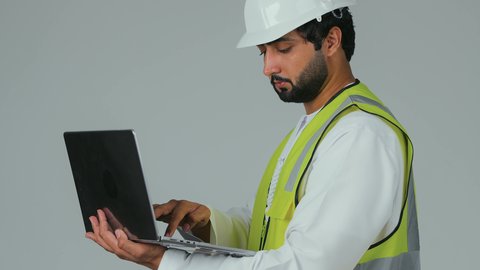 إنجاز مهام العمل عن بعد، النقر على لوحة المفاتيح، استخدام الأجهزة التقنية الحديثة، بورتريه لمهندس عربي خليجي امارتي يرتدي الكندورة والخوذة وسترة الحماية يستخدم الحاسوب المحمول، متابعة المشاريع المعمارية والهندسية، ايماءات الاندماج و التركيز، خلفية بيضاء