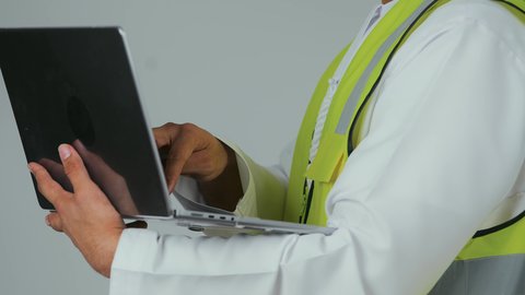 النقر على لوحة المفاتيح، إنجاز مهام العمل عن بعد، استخدام الأجهزة التقنية الحديثة، بورتريه مقرب ليد لمهندس عربي خليجي امارتي يرتدي الكندورة وسترة الحماية يستخدم الحاسوب المحمول، متابعة المشاريع المعمارية والهندسية، خلفية بيضاء