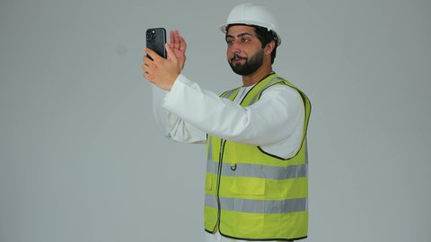 دمج التكنولوجيا المتطورة بالهندسة، رفع كف بتعابير التحية و السلام،  اجراء مكالمة فيديو عبر الجوال, بورتريه لمهندس عربي خليجي امارتي يرتدي الكندورة وسترة الحماية يقوم بمتابعة سير الأعمال عن بعد عبر الهاتف المحمول، خلفية بيضاء