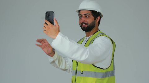 رفع كف بتعابير التحية و السلام،  اجراء مكالمة فيديو عبر الجوال, بورتريه لمهندس عربي خليجي امارتي يرتدي الكندورة وسترة الحماية يقوم بمتابعة سير الأعمال عن بعد عبر الهاتف المحمول، دمج التكنولوجيا المتطورة بالهندسة، خلفية بيضاء