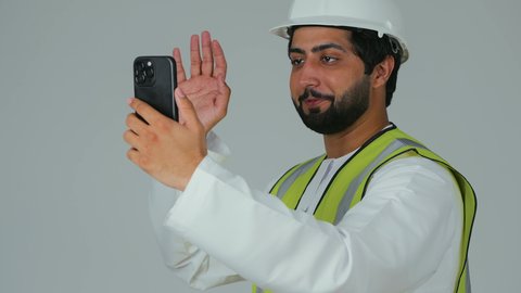 اجراء مكالمة فيديو عبر الجوال، رفع كف بتعابير التحية و السلام, بورتريه لمهندس عربي خليجي امارتي يرتدي الكندورة وسترة الحماية يقوم بمتابعة سير الأعمال عن بعد عبر الهاتف المحمول، دمج التكنولوجيا المتطورة بالهندسة، خلفية بيضاء