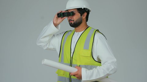 إدارة ومراقبة المشاريع الإنشائية، رؤية الأشياء البعيدة، بورتريه لمهندس عربي خليجي إماراتي يرتدي الكندورة والخوذة وسترة الحماية ينظر من خلال المنظار ثنائي العينين إلى شيء ما يحمل بيده مخطط، المهن والوظائف الشبابية، خلفية بيضاء