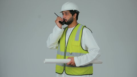 الإشراف على أعمال البناء والتعمير، بورتريه لمهندس عربي خليجي إماراتي يرتدي الكندورة والخوذة وسترة الحماية يحمل جهاز اتصال لاسلكي يمسك بيده مخطط، إعطاء أوامر خاصة بالعمل، المهن والوظائف الشبابية في الإمارات العربية المتحدة، خلفية بيضاء