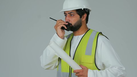 مراقبة وتنفيذ المشاريع الهندسية الإنشائية، بورتريه لمهندس عربي خليجي إماراتي يرتدي الكندورة والخوذة وسترة الحماية يحمل جهاز اتصال لاسلكي يمسك بيده مخطط، إعطاء أوامر خاصة بالعمل، المهن والوظائف الشبابية في الإمارات العربية المتحدة، خلفية بيضاء