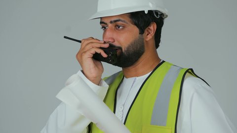 المتابعة والإشراف على المشاريع الهندسية الإعمارية، بورتريه لمهندس عربي خليجي إماراتي يرتدي الكندورة والخوذة وسترة الحماية يحمل جهاز اتصال لاسلكي يمسك بيده مخطط، إعطاء أوامر خاصة بالعمل، المهن والوظائف الشبابية في الإمارات العربية المتحدة، خلفية بيضاء