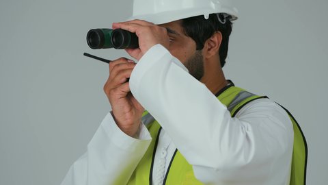 إعطاء أوامر خاصة بالعمل، رؤية الأشياء البعيدة، متابعة سير المشاريع الهندسية، مهن ووظائف شبابية، بورتريه مقرب لمهندس عربي خليجي إماراتي يرتدي الكندورة والخوذة وسترة الحماية يحمل جهاز اتصال لاسلكي ينظر من خلال المنظار ثنائي العينين، خلفية بيضاء