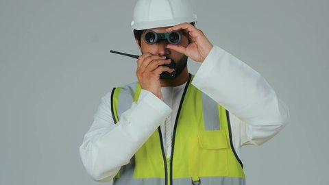 متابعة سير المشاريع الهندسية، مهن ووظائف شبابية، رؤية الأشياء البعيدة، بورتريه لمهندس عربي خليجي إماراتي يرتدي الكندورة والخوذة وسترة الحماية يحمل جهاز اتصال لاسلكي ينظر من خلال المنظار ثنائي العينين، إعطاء أوامر خاصة بالعمل، خلفية بيضاء
