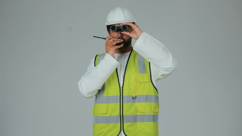 مهن ووظائف شبابية، متابعة سير المشاريع الهندسية، رؤية الأشياء البعيدة، بورتريه لمهندس عربي خليجي إماراتي يرتدي الكندورة والخوذة وسترة الحماية يحمل جهاز اتصال لاسلكي ينظر من خلال المنظار ثنائي العينين، إعطاء أوامر خاصة بالعمل، خلفية بيضاء