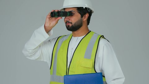 توسيع مجال الرؤية، التخطيط والبحث للمستقبل، الإشراف على المشاريع الهندسية، القطاع الهندسي في الإمارات، بورتريه مقرب لمهندس عربي خليجي إماراتي يرتدي الكندورة والخوذة وسترة الحماية ينظر عبر منظار ثنائي العينين يحمل بيده ملف ازرق خاص بالعمل، خلفية بيضاء