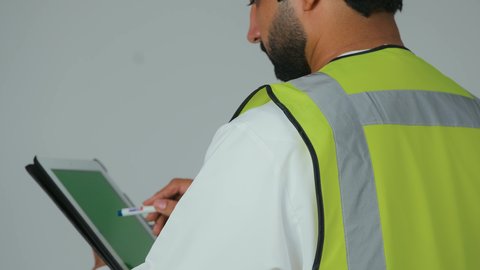 كتابة آخر المستجدات حول المشروع، التأكد من سير الأعمال بالشكل المطلوب والصحيح، تدوين و تسجيل الملاحظات، بورتريه  مقرب من الخلف لمهندس عربي خليجي إماراتي يرتدي الكندورة والخوذة وسترة الحماية يقوم باستخدام الجهاز اللوحي، الأجهزة التقنية الحديثة، خلفية بيضاء
