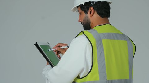 التأكد من سير الأعمال بالشكل المطلوب والصحيح، كتابة آخر المستجدات حول المشروع، تدوين و تسجيل الملاحظات، بورتريه من الخلف لمهندس عربي خليجي إماراتي يرتدي الكندورة والخوذة وسترة الحماية يقوم باستخدام الجهاز اللوحي، الأجهزة التقنية الحديثة، خلفية بيضاء