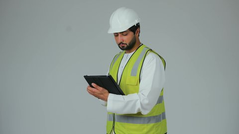 Engineering project management, completing remote work tasks, modern technical devices, ensuring work is progressing as required, a portrait of an Arab Gulf Emirati engineer wearing a kandura, helmet, and safety vest using a tablet, white background.