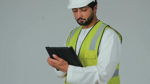 الأجهزة التقنية الحديثة، إنجاز مهام العمل عن بعد، التأكد من سير الأعمال بالشكل المطلوب، بورتريه لمهندس عربي خليجي إماراتي يرتدي الكندورة والخوذة وسترة الحماية يقوم باستخدام الجهاز اللوحي، تدوين و تسجيل الملاحظات، خلفية بيضاء