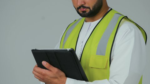 إنجاز مهام العمل عن بعد، الأجهزة التقنية الحديثة، التأكد من سير الأعمال بالشكل المطلوب، بورتريه لمهندس عربي خليجي إماراتي يرتدي الكندورة والخوذة وسترة الحماية يقوم باستخدام الجهاز اللوحي، تدوين و تسجيل الملاحظات، خلفية بيضاء