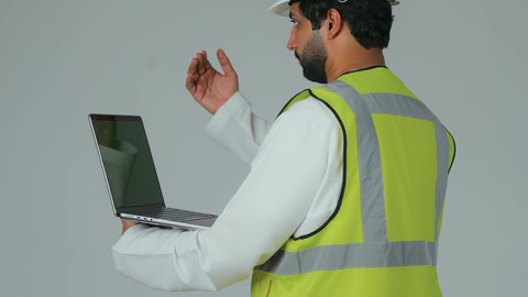 Giving specific work orders, supervising and following up on work, engineering professions and jobs, managing architectural engineering projects, a portrait from behind of an Arab Gulf Emirati engineer wearing a helmet and safety vest holding a laptop, white background.