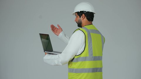 الإشراف ومتابعة العمل ، إعطاء أوامر خاصة بالعمل ، مهن و وظائف هندسية ، ادارة المشاريع المعمارية الهندسية، بورترية من الخلف لمهندس عربي خليجي اماراتي يرتدي خوذة و ستره الحماية  يحمل الحاسب المحمول، خلفية بيضاء