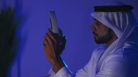 مفهوم الإصغاء والاستماع، الاستفادة من التكنولوجيا الحديثة، إستخدام تقنية الهيدفون، قضاء أوقات ممتعة بسماع الاغاني والمقاطع الصوتية، تصوير مقرب لرجل عربي خيجي اماراتي يرتدي الكندورة والغترة يقوم بوضع سماعة الرأس