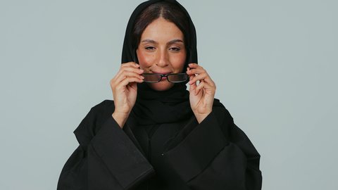 الاهتمام بالمظهر الخارجي، ارتداء النظارات الخاصة بضعف النظر، بورتريه مقرب لامرأة عربية خليجية سعودية محجبة ترتدي العباءة و الحجاب تمسك بيدها نظارة طبية تقف بايدي متقاطعة، تحسين الرؤية ومحاولة تعديل البصر، خلفية رمادي