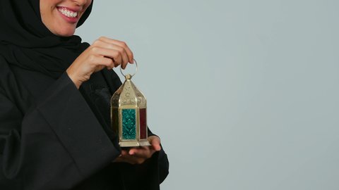 المناسبات والأعياد الدينية، الإستعداد لقدوم شهر رمضان، العبادة والتقرب إلى الله، أجواء السكينة والروحانية، بورتريه مقرب من الجانب لإمراة عربية خليجية سعودية محجبة ترتدي العباءة تحمل بيدها فانوس معدني،الاشارة بإصبع السبابة الى شيء ما، خلفية رمادي