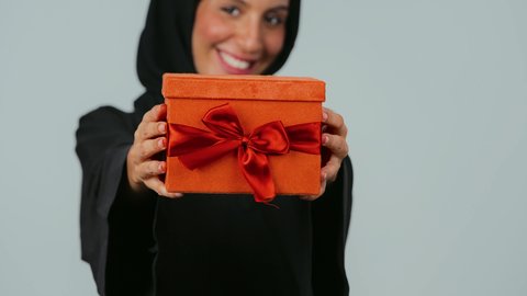 السعادة بتقديم الهدايا والتهنئات، ايماءات الفرح عند الحصول على الهدايا، التعبير عن المحبة والتقدير، أعياد ومناسبات سعيدة, بورتريه لامرأة عربية خليجية سعودية ترتدي العباءة السوداء والحجاب تحمل بيديها علبة مغلفة، خلفية رمادي