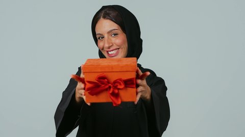 Gestures of joy when receiving gifts, expressions of love and appreciation, happiness in giving gifts and congratulations, happy holidays and occasions, a portrait of a Saudi Gulf Arab woman wearing a black abaya and hijab holding a wrapped box in her hands, gray background.