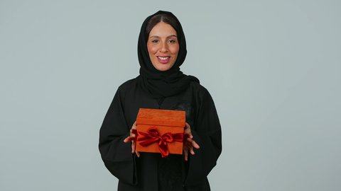 التعبير عن المحبة والتقدير، ايماءات الفرح عند الحصول على الهدايا، السعادة بتقديم الهدايا والتهنئات، أعياد ومناسبات سعيدة, بورتريه لامرأة عربية خليجية سعودية ترتدي العباءة السوداء والحجاب تحمل بيديها علبة مغلفة، خلفية رمادي