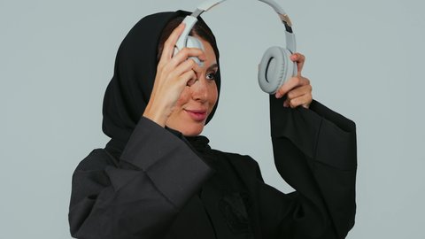 الأجهزة التقنية الحديثة، الاستمتاع بسماع الموسيقى المفضلة، قضاء أوقات ممتعة بسماع الأغاني، بورتريه مقرب لامرأة عربية خليجية سعودية ترتدي الحجاب والعباءة تضع سماعات الرأس، ايماءات الاندماج و السرور، خلفية رمادي