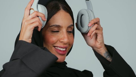 Enjoying listening to favorite music, modern tech devices, spending enjoyable times listening to songs, a close-up portrait of a Saudi Gulf Arab woman wearing a hijab and abaya with headphones on, gestures of immersion and joy, gray background.