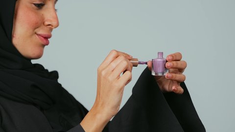 مفهوم العناية بالأظافر, الاهتمام بالأناقة والمظهر الخارجي, التسويق لمنتجات ومستحضرات التجميل, بورتريه مقرب من الجانب لامرأة عربية خليجية سعودي محجبة ترتدي العباءة تمسك بيدها علبة طلاء الأظافر, خلفية رمادي