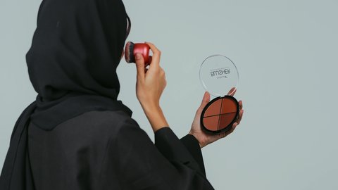 حب وضع المكياج وتنسيقه، صالون نسائي راقي، أدوات تجميل نسائيه عصريه، التسويق لمنتجات التجميل, بورتريه مقرب من الخلف لفتاه عربيه سعودية خليجيه ترتدي الحجاب و العباءة تقوم بوضع توريد الخدود, خلفيه رمادي