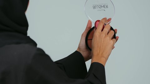 صالون نسائي راقي، أدوات تجميل نسائيه عصريه، التسويق لمنتجات التجميل, بورتريه مقرب من الخلف لفتاه عربيه سعودية خليجيه ترتدي الحجاب و العباءة تقوم بوضع توريد الخدود, مفهوم الانوثه والاهتمام بالمظهر الخارجي, مكياج وميكاب تتوريال, خلفيه رمادي