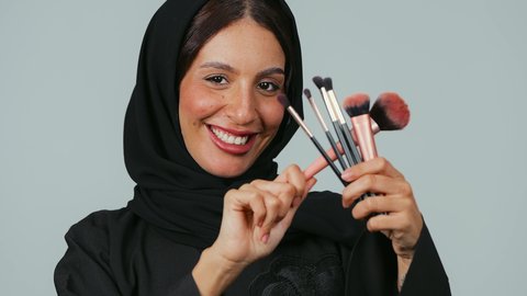 The passion for decorating the face with makeup, looking at the camera with gestures of happiness and joy, caring for and taking care of the skin and beauty, the concept of adornment and beautification, a close-up portrait of a Saudi Arabian Gulf woman wearing a hijab and an abaya holding makeup brushes in her hands, gray background.