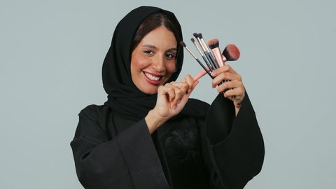 Love for makeup experimentation to highlight beauty, looking at the camera with gestures of happiness and joy, caring for skin and beauty, the concept of adornment and beautification, a close-up portrait of a Saudi Arabian Gulf woman wearing a hijab and an abaya holding makeup brushes in her hands, gray background.