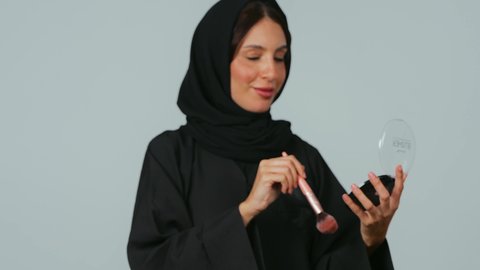 النظر إلى الكاميرا بإيماءات السعادة والسرور، الاهتمام والعناية بالبشرة والجمال، مفهوم التزين والتجمل, الشغف بوضع المكياج, بورتريه لامرأة عربية خليجية سعودية محجبة ترتدي العباءة تمسك بيديها علبة وفرشاة أحمر الخدود, خلفية رمادي