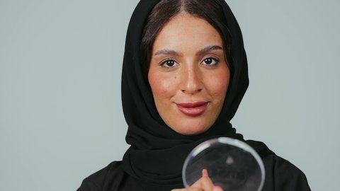 الأنوثة والاهتمام بالجمال والبشرة والمظهر الخارجي, مفهوم الدعاية والإعلان، خبيرة لمستحضرات التجميل النسائية، بورتريه مقرب لإمرأة عربية خليجية سعودية ترتدي العباءة والحجاب تقوم بوضع أحمر الخدود، التسويق لمنتجات الميك اب، خلفية رمادي