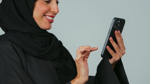 Gestures of joy and happiness, browsing social media applications and programs, chatting with family and friends, a close-up side portrait of a smiling Saudi Gulf Arab woman wearing an abaya and hijab holding a mobile phone in her hand, using modern and advanced technological devices, gray background.