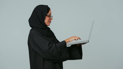 إدارة المهام عن بعد، النقر على لوحة المفاتيح، انجاز العمل باستخدام التقنيات الحديثه، استخدام الأجهزة التقنية الحديثة والمتطورة, بورتريه من الجانب لسيده عربية خليجية سعوديه محجبة ترتدي العباءة و الحجاب و النظارات الطبية  تعمل على جهاز الحاسوب المحمول، خلفية رمادي