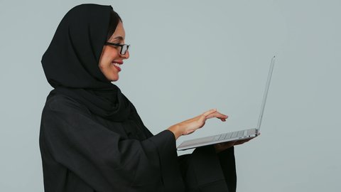 ممارسة العمل عن بعد، النقر على لوحة المفاتيح، انجاز المهام باستخدام التقنيات الحديثه، استخدام الأجهزة التقنية الحديثة والمتطورة, بورتريه من الجانب لسيده عربية خليجية سعوديه محجبة ترتدي العباءة و الحجاب و النظارات الطبية  تعمل على جهاز الحاسوب المحمول، خلفية رمادي