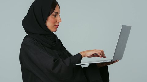 مفهوم العمل عن بعد، استخدام الأجهزة التقنية الحديثة والمتطورة, بورتريه من الجانب لسيده عربية خليجية سعوديه محجبة ترتدي العباءة و الحجاب تعمل على جهاز الحاسوب المحمول, أنجاز المهام باستخدام التقنيات الحديثه،النقر على لوحة المفاتيح، خلفية رمادي