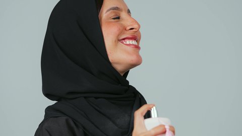 التطيب بالعطور الفاخرة، التسويق لمنتجات الروائح الوطنية في المملكة السعودية، مفهوم الأناقة والاهتمام بالمظهر الخارجي، استخدام الزيوت العطرية الطبيعية، بورتريه مقرب لشابة عربية خليجية سعودية محجبة ترتدي عباءة  سوداء تمسك بيدها زجاجة وتقوم برش العطر, خلفية رمادي