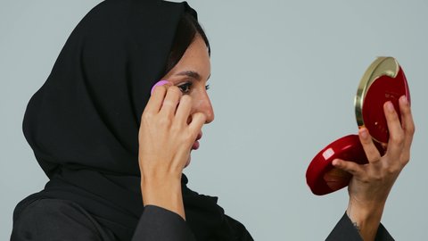 الشغف بوضع المكياج، الاهتمام بالفاشن ومواكبة الموضة، الوجوه الإعلانية التسويقية لبراندات الميكاب، بورتريه مقرب من الجانب لشابة عربية خليجية سعودية محجبة ترتدي العباءة الملونة تمسك بيدها مستحضرات التجميل وتقوم بتجربتها بإيماءات السرور، خلفية رمادي