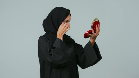 الوجوه الإعلانية التسويقية لبراندات الميكاب، الاهتمام بالفاشن ومواكبة الموضة، الشغف بوضع المكياج، بورتريه من الجانب لشابة عربية خليجية سعودية محجبة ترتدي العباءة الملونة تمسك بيدها مستحضرات التجميل وتقوم بتجربتها بإيماءات السرور، خلفية رمادي